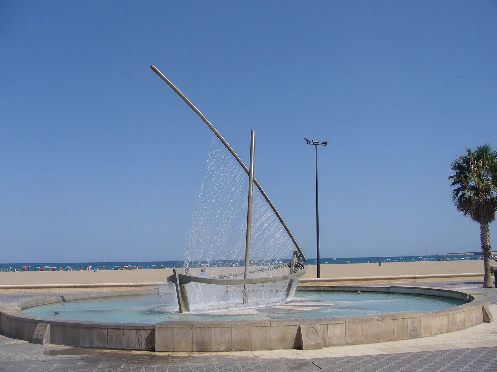 Segelförmiger Brunnen, dessen Wasser in ein rundes Becken neben einem Sandstrand fällt