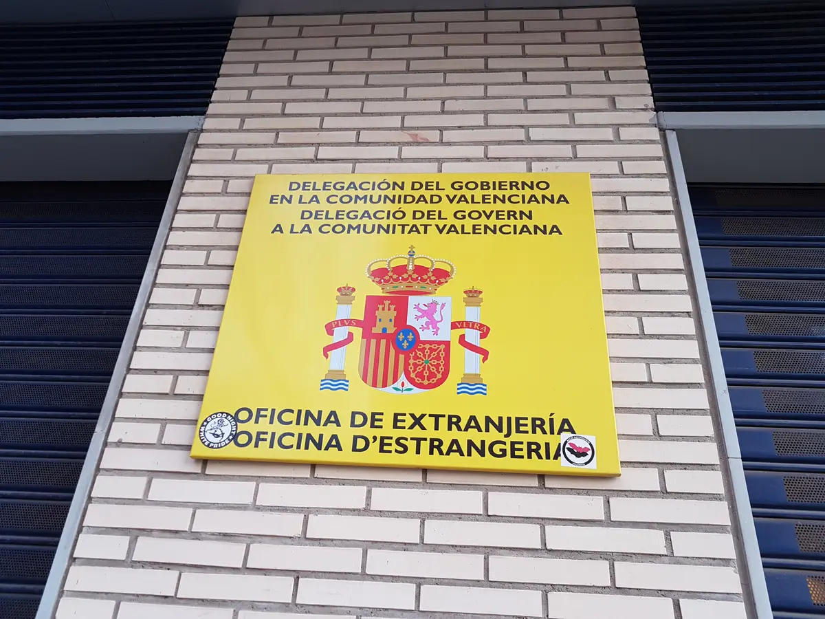Gelbes Schild der Oficina de Extranjería der Regierungsdelegation an einer Backsteinmauer.