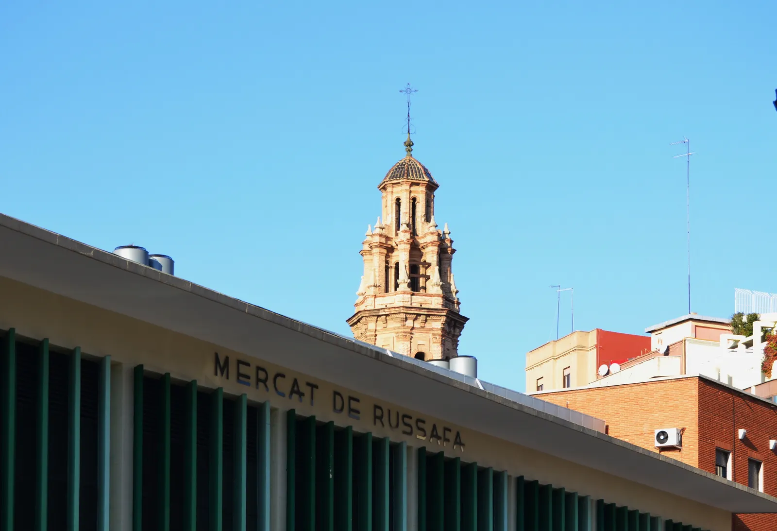 Dachkante des Mercat de Russafa mit dem Namen an der Fassade und einem Kirchturm dahinter.