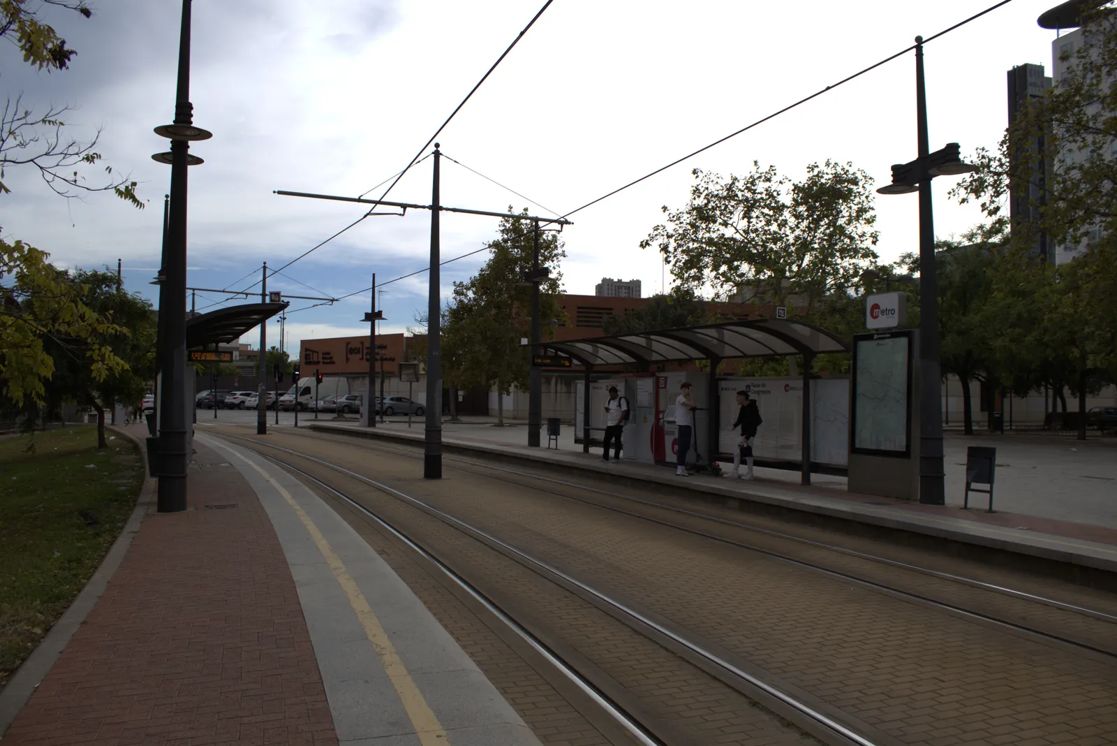 Überdachte Tramhaltestelle neben einem baumgesäumten Gleis, Fahrgäste warten auf dem Bahnsteig