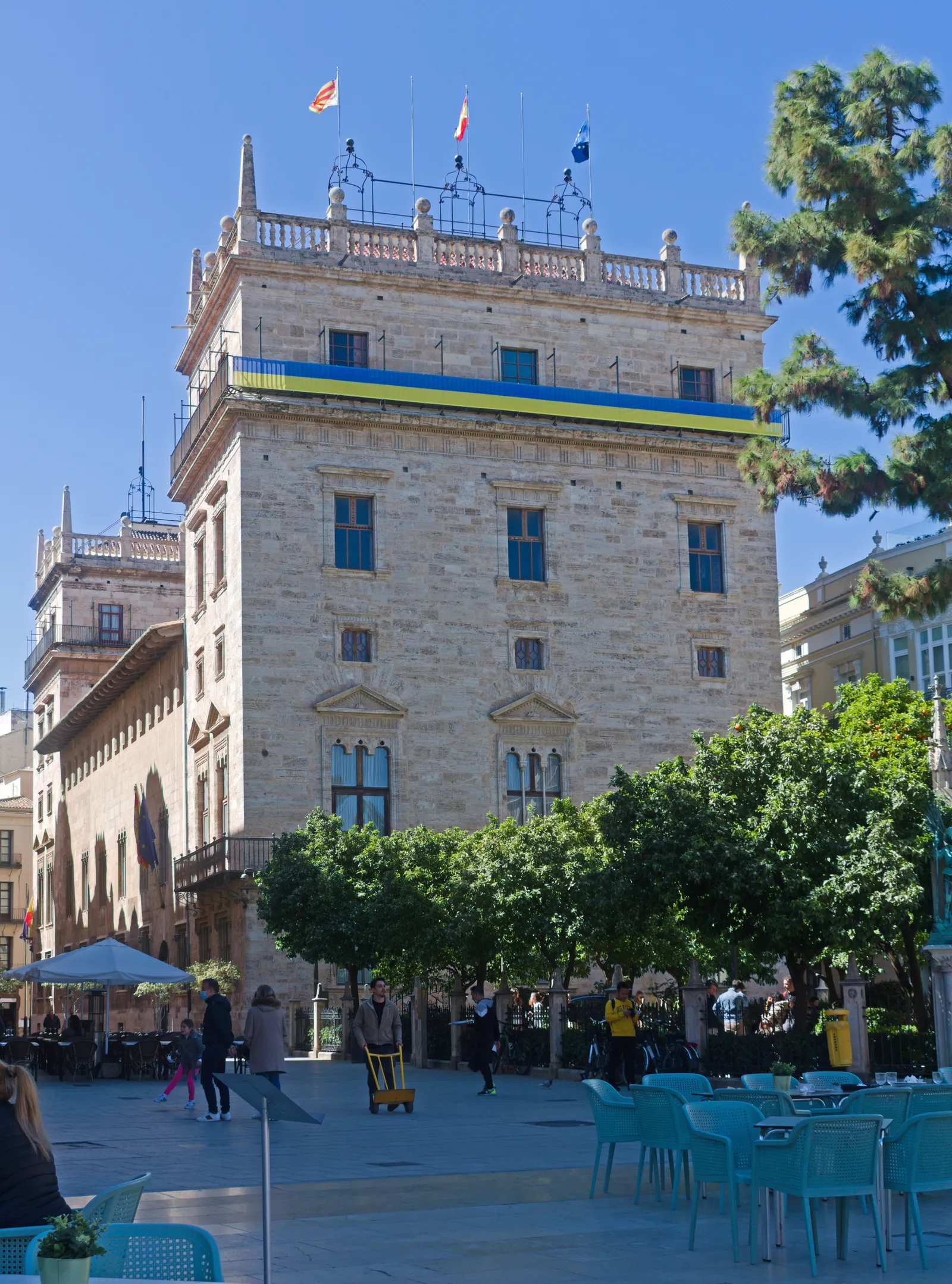 Historischer Palast mit Steinturm und Reihen von Bogenfenstern, davor ein Straßencafé und Bäume.