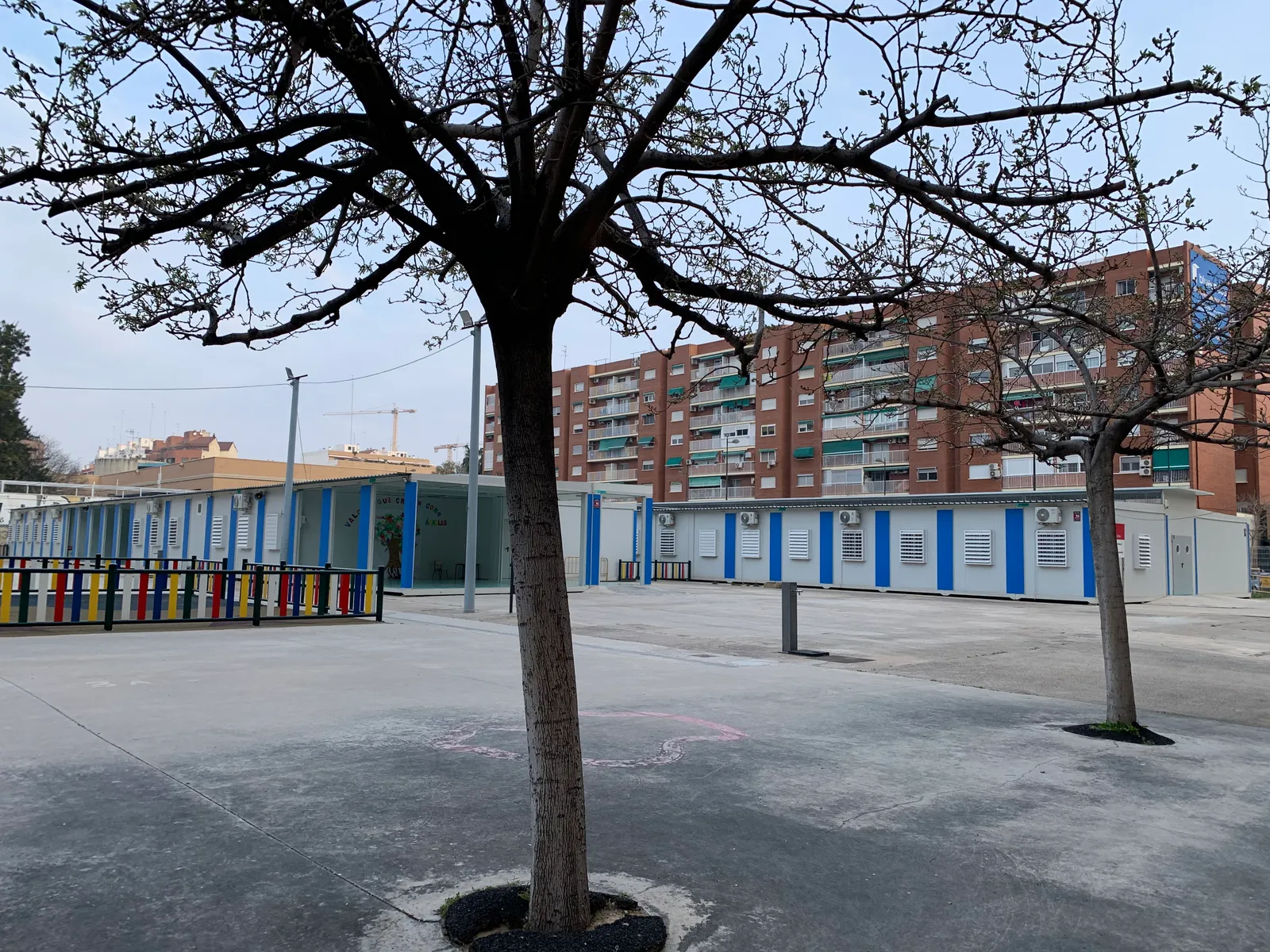 Leerer Hof einer Grundschule in Valencia mit kahlen Bäumen, niedrigen Klassenzimmerblöcken mit blauen Streifen und einem Backstein-Wohnhaus dahinter.